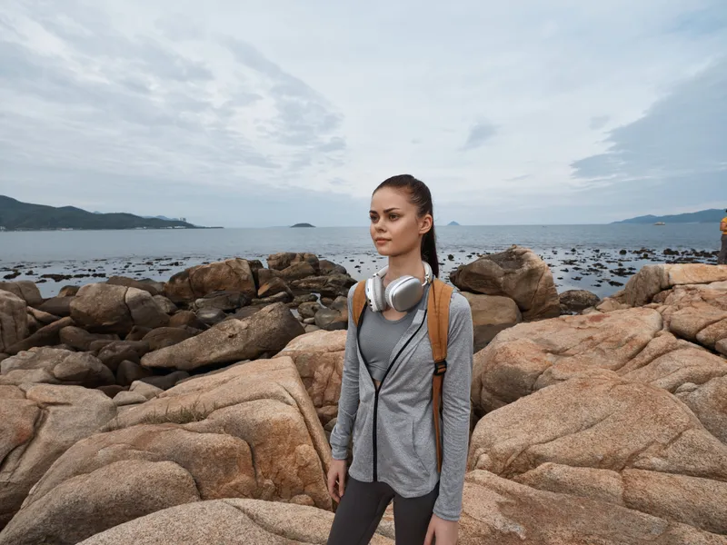 Young woman in activewear stands on rocky beach, headphones around neck, ocean backdrop, showcasing a confident vibe and healthy lifestyle