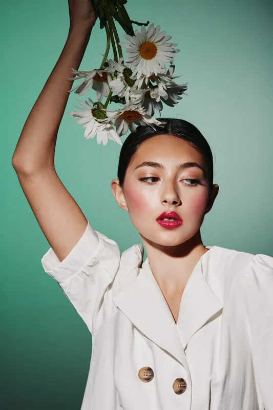 Young stylish woman with red lipstick holds daisies above her head against green background. Fashion creative with glossy makeup and elegant white outfit.
