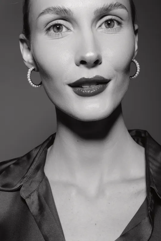 Close-up portrait of a fair-skinned Caucasian woman with short hair and hoop earrings wearing a dark blouse. Studio lighting highlights facial features and expression.