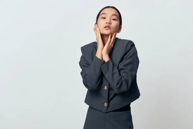 Woman posing with hands near face in stylish dark suit against white background. Portrait of confident female model with minimal makeup and neat hair for fashion and beauty concept.