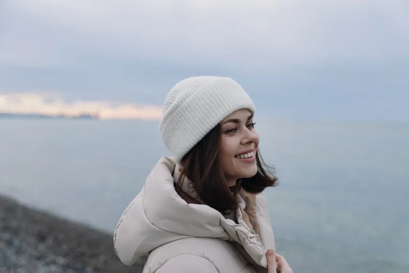 Woman portrait in a calm coastal setting. Outdoor winter scene with a smiling face, soft white beanie and warm jacket by the sea, gentle breeze and peaceful horizon.