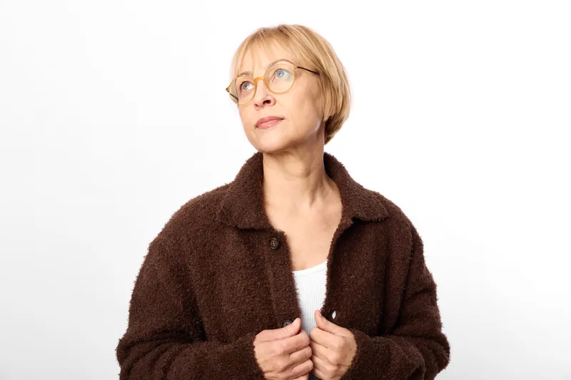 Senior woman with glasses and short blonde hair, wearing a brown cozy jacket, portrait against a white background, calm thoughtful mood, studio lighting