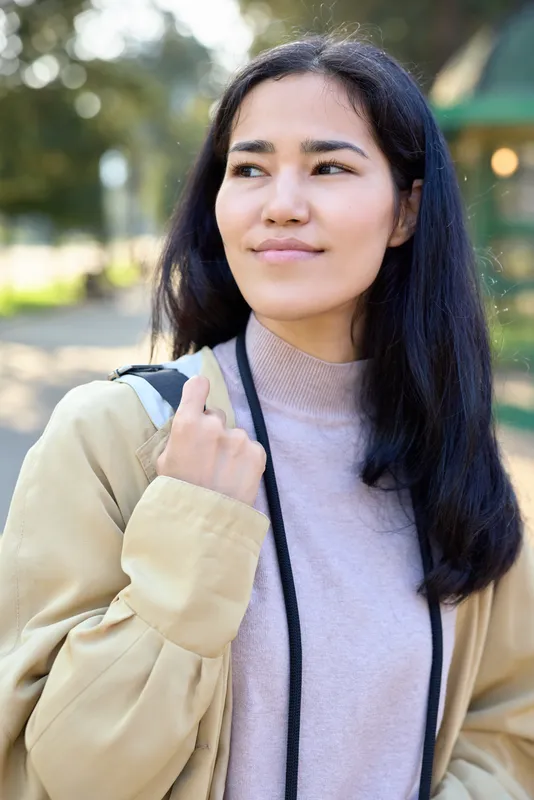 Woman in beige jacket and lavender sweater standing outdoors in a park, thoughtful expression, soft sunlight, casual style, natural background, lifestyle moment in daytime calm setting