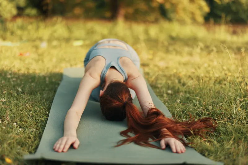 A serene young woman practicing yoga in a green park, showcasing a flexible pose, with red hair flowing on a yoga mat, surrounded by lush greenery
