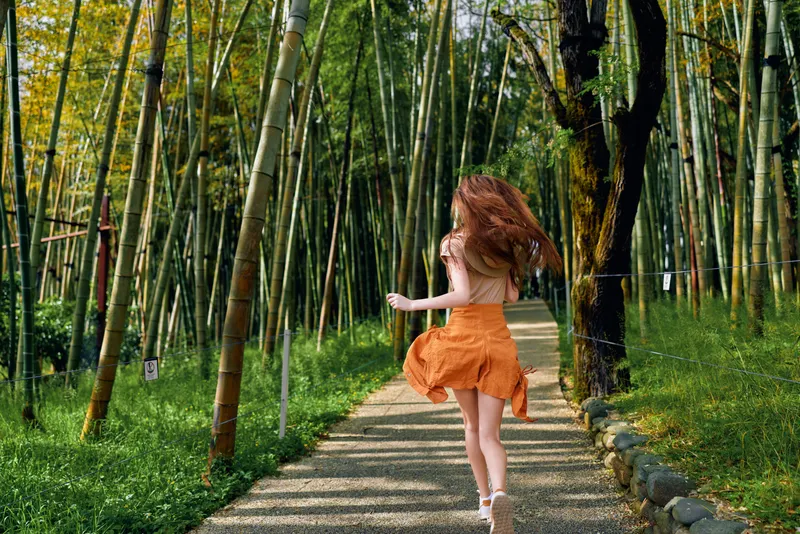 Woman running through bamboo forest on a gravel path in orange dress, back view of young girl enjoying nature, movement and sunlight in serene outdoor scenery.