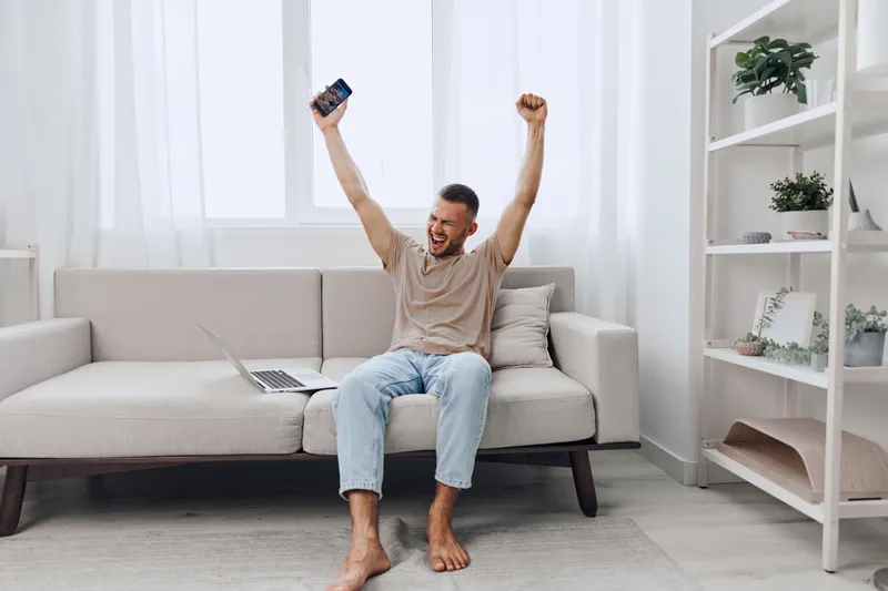 Excited man celebrating success while sitting on a cozy couch, wearing casual clothing, surrounded by minimalistic decor, and holding a smartphone.