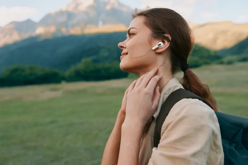 Young woman with brunette hair listens to wireless earbuds while strolling through a sunlit grassy field with distant mountains, exuding a relaxed outdoor mood.