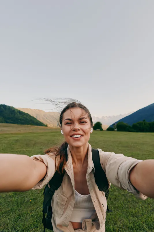 Young woman outdoors takes a selfie while hiking through a lush green valley, smiling with wind in her hair and a backpack on her shoulders, bright summer light and open landscape