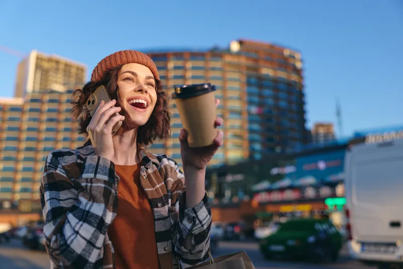 Woman coffee phone city smile candid golden hour young woman holding takeaway cup and talking on smartphone in urban street, authentic candid lifestyle, mindful living and emotional storytelling.