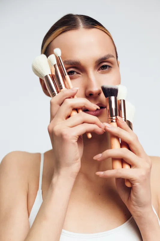 A young woman holds makeup brushes near her face, focusing on applying cosmetics in a bright studio, conveying concentration and artistry during a beauty shoot.