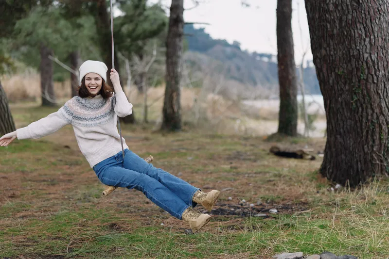 Woman on a swing in a park, bundled in a warm sweater and beanie, enjoying outdoor play among trees and grass on a crisp day.