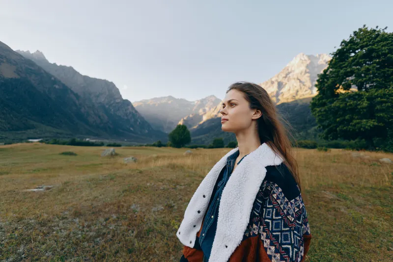 Woman portrait in mountains meadow, nature outdoors and travel landscape setting with thoughtful expression, cozy patterned coat and wide valley view at golden hour.