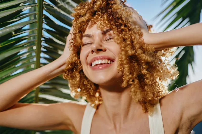 Smiling woman with curly hair enjoys tropical sun under green palm leaves, radiating happiness and natural beauty in bright daylight outdoors.