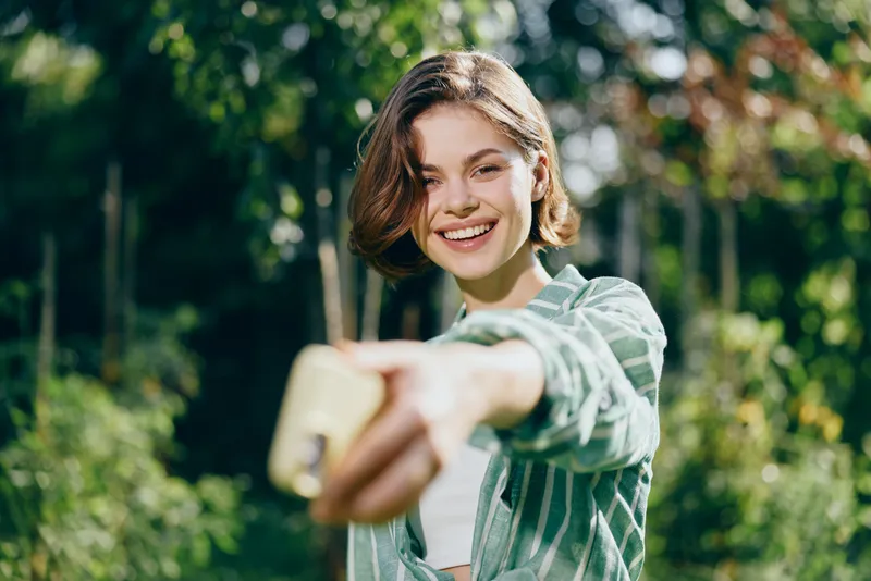 Cheerful young woman outdoors, extending arm to take a selfie with a bright smile in a sunny park setting, casual striped shirt.