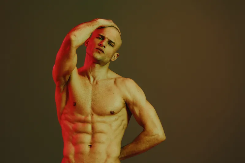 Muscular man posing gracefully against a gradient background, showcasing strength and confidence. The lighting emphasizes contours and adds a striking visual element.