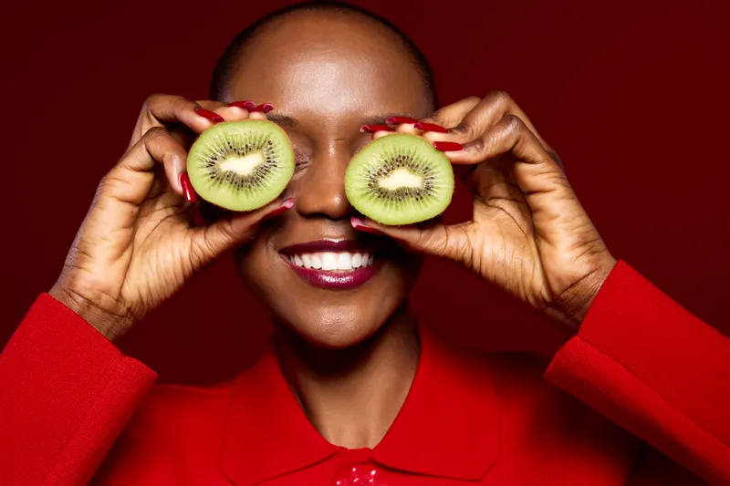 Smiling African woman with dark skin holds two kiwi halves in front of eyes. She wears a red top and has red nails. Bright studio lighting highlights fresh fruit and joyful mood.
