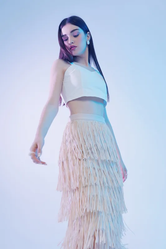 Young woman in a white crop top and fringed skirt poses gracefully against a soft gradient background. Concept of beauty, fashion, and modern style with elegant lighting.