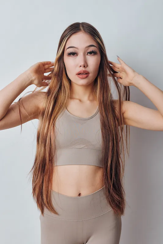 Woman with long hair in beige sportswear posing with hands near head against white background. Fitness model, beauty and style with confident expression in studio lighting.
