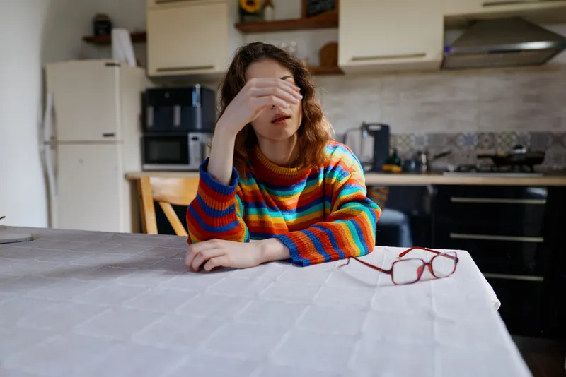 Woman with a colorful striped sweater rests her head on her hand at a kitchen table, creating a calm home lifestyle moment that suggests fatigue, focus, and everyday weariness.