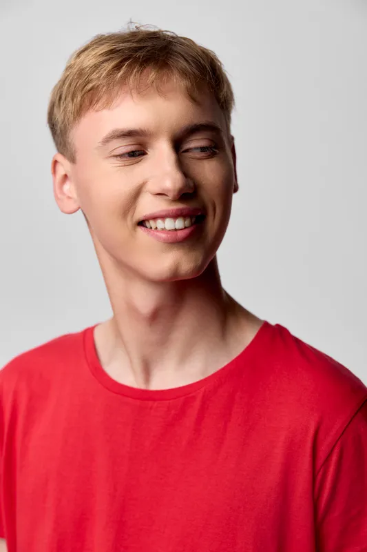 Smiling young man in a red shirt looks to the side with a warm expression, casual studio shot, clean background, natural lighting, relaxed mood, suitable for lifestyle, marketing and promo use