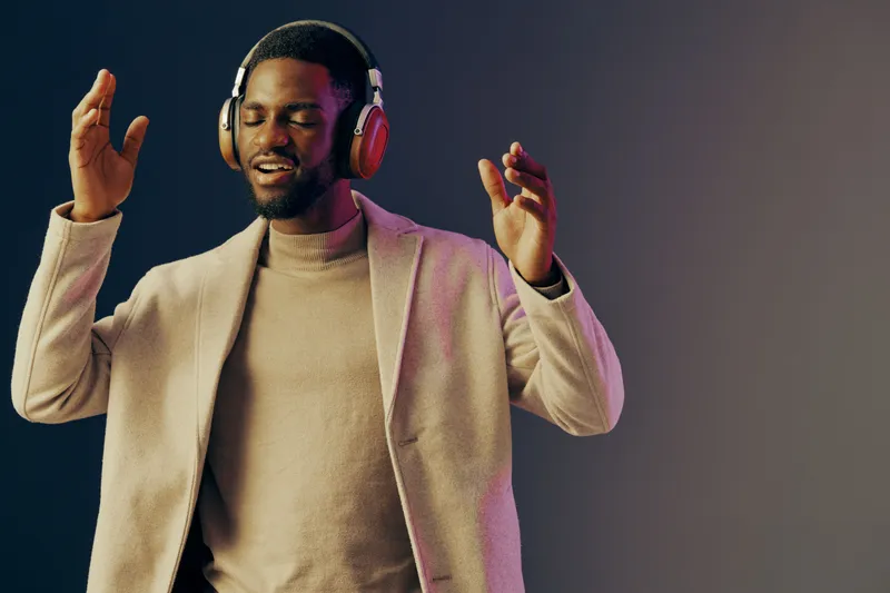 Young man enjoying music with headphones, wearing a stylish coat, smiling joyfully against a gradient background, expressing happiness and relaxation