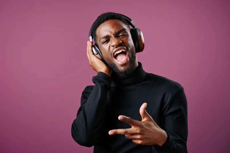Young African man enjoying music with headphones, wearing a black turtleneck, expressing joy and passion against a pink background