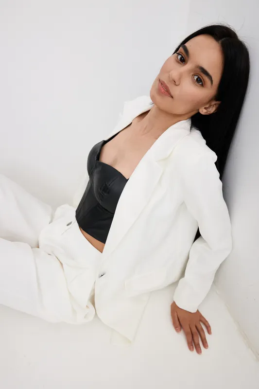 Young woman in white suit and black top sitting on floor against wall, dark hair, natural makeup, stylish casual outfit, indoor minimalistic setting.