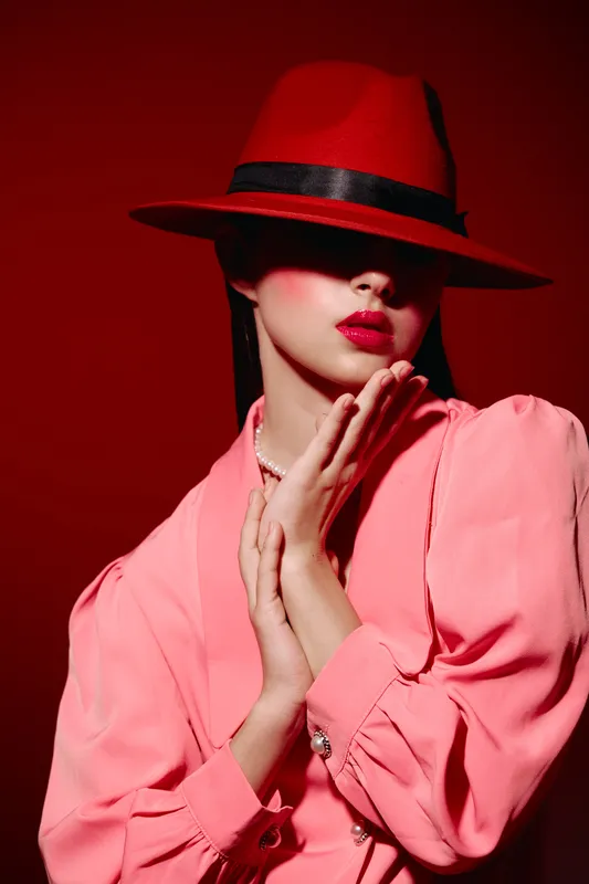 Stylish woman wearing a bold red hat partially covering her face poses with hands near chin. She wears pink blouse and bright lipstick against intense red background, fashion beauty concept.