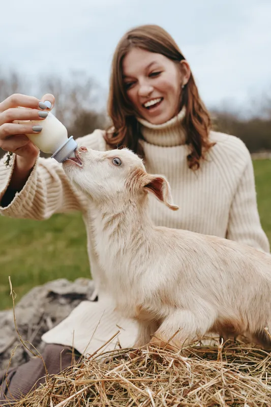 A smiling woman bottle feeds a baby goat outdoors on a farm, capturing a warm lifestyle moment of care, bonding, and tenderness between caretaker and animal amid hay and pasture