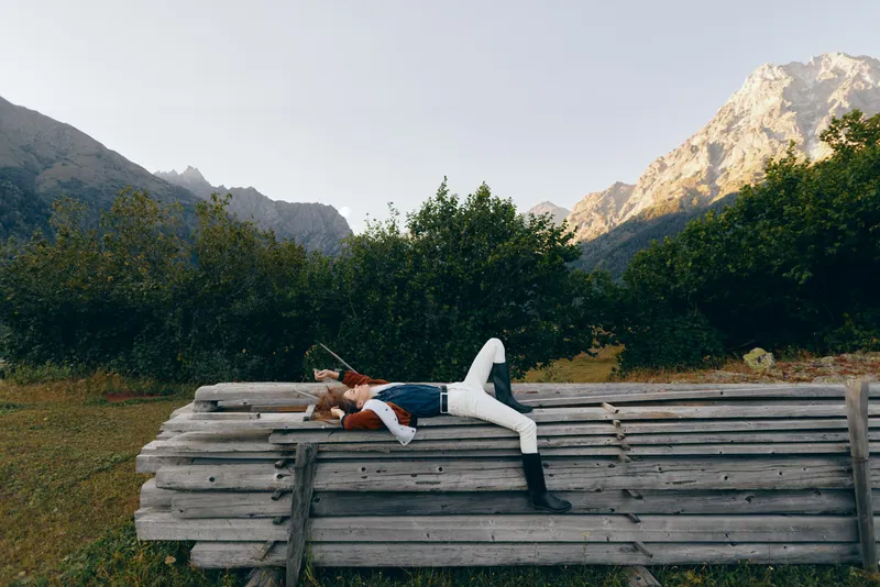woman lying on wooden bench in mountain nature scene with forest backdrop and meadow, relaxed outdoors lifestyle portrait, casual fashion and peaceful resting in scenic travel escape