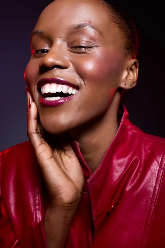 Close-up of a smiling black woman with dark skin wearing a red leather jacket. She touches her face gently, showing confidence and joy. Studio portrait with dramatic lighting.