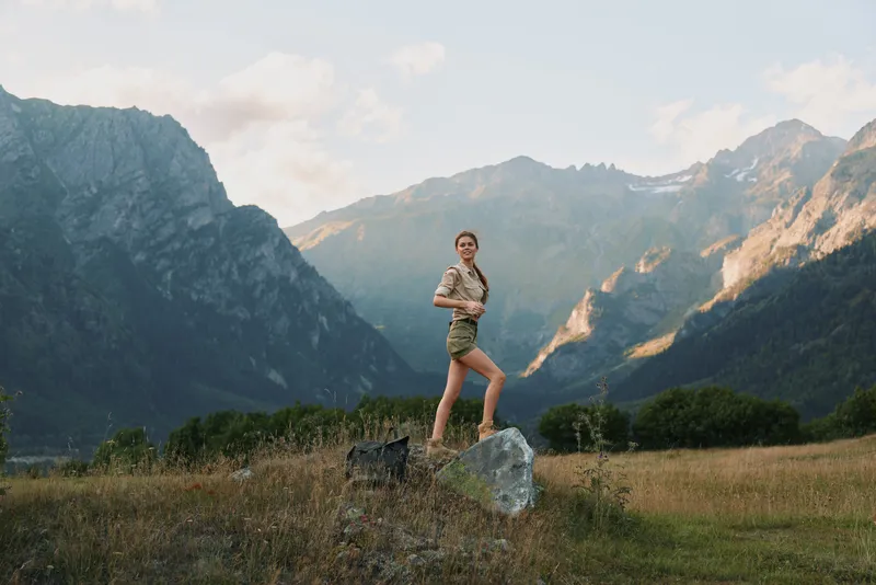 Young woman in casual outfit stands confidently on a rock in a mountain valley during golden hour, enjoying nature and fresh air with a relaxed expression.