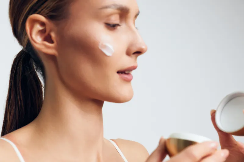 Woman in profile applying face cream on her cheek, skincare care self wellness concept, beauty routine with moisturizer, soft studio light, closeup on white background