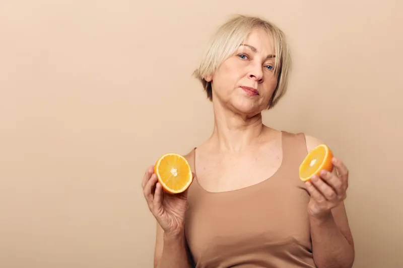 Mature woman with short blonde hair holds two halves of fresh orange fruit against beige background. Concept for healthy lifestyle, nutrition, vitamins, and natural wellness.