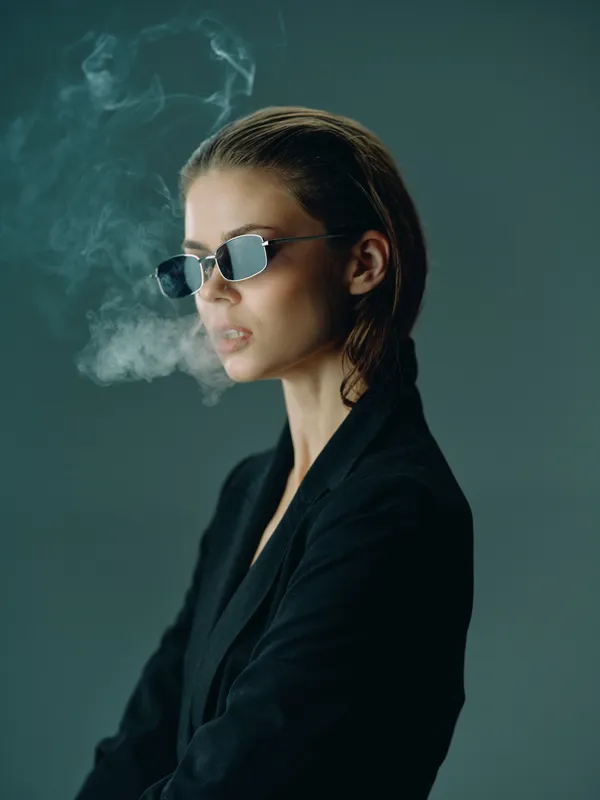 Stylish woman wearing sunglasses and black jacket exhaling smoke in moody lighting with modern aesthetic and cool attitude.