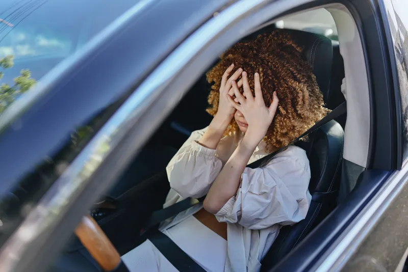 Stressed woman covering face with hands inside car wearing seatbelt with curly hair and white blouse, expressing frustration or anxiety during daytime drive.