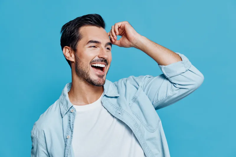 Handsome smiling man with stylish hair wearing a light blue denim shirt, posing joyfully against a vibrant blue background. Cheerful expression concept.
