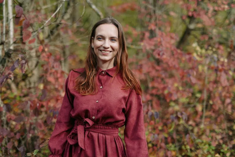 Authentic portrait of a smiling woman in a burgundy dress standing in a natural outdoor setting, conveying genuine presence, honest emotion, and everyday beauty.