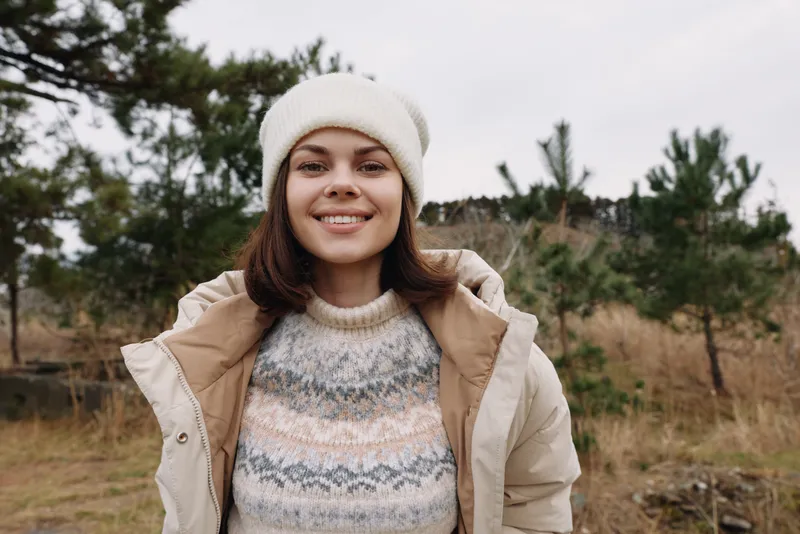 Woman outdoors in autumn attire with a beige jacket, knit sweater and beanie, smiling against a natural landscape of trees and fields for a fresh lifestyle shot, capturing cozy style and outdoor mood.