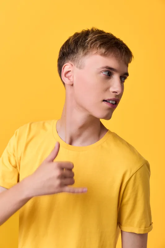 Male subject in a bright yellow tee making a hand gesture with thumb and pinky extended against a bold yellow backdrop, casual style, energetic and confident mood, modern lifestyle