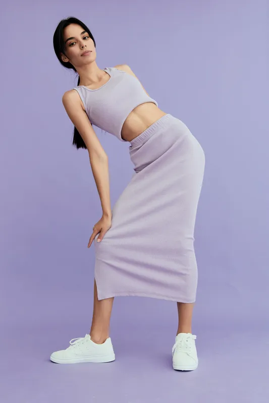 Young woman in a lilac crop top and skirt posing confidently against a purple background, wearing white sneakers, showcasing modern casual fashion style.