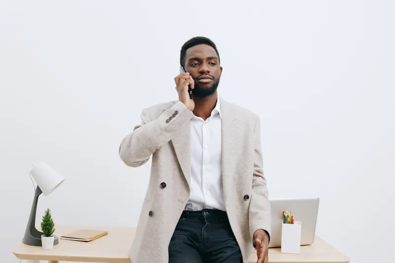 Stylish young man with dark skin in smart casual outfit talking on mobile phone while standing in modern office environment. Business professional, communication, technology, workspace, confident pose