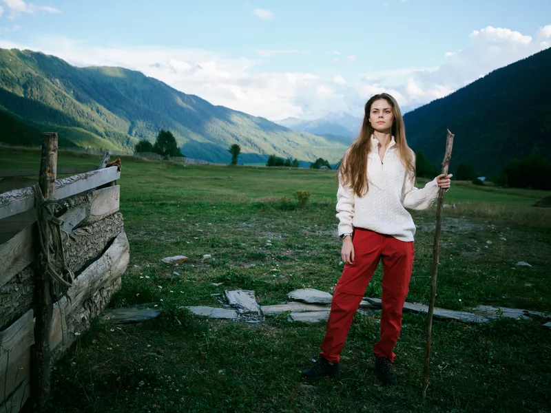 Woman stands in a green valley with distant mountains, holding a wooden staff, wearing a white sweater and red pants, embodying outdoor adventure, rustic countryside, and serene landscape