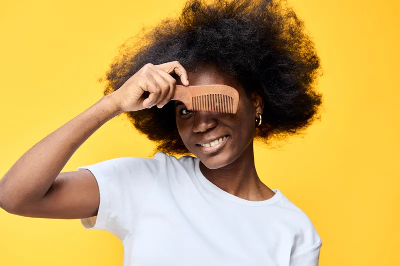 Happy Black woman with curly hair holding a wooden comb, smiling against a vibrant yellow background, representing confidence and natural beauty