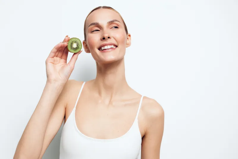 Young woman with blonde hair smiles while holding a half of fresh kiwi fruit near her face against a white background.
