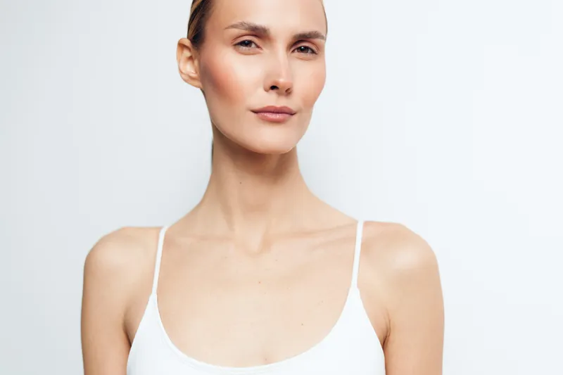 Young woman with blonde hair and minimal makeup poses confidently in a white tank top against a plain white background showing natural skin texture and calm expression.