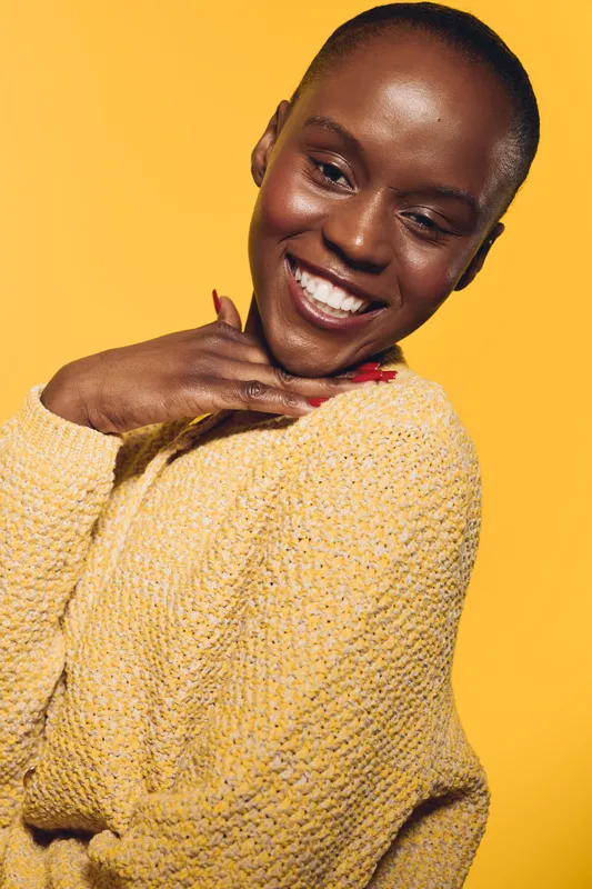 Happy African woman with dark skin smiles brightly, wearing a yellow sweater against yellow background. Portrait of joyful black female model expressing positivity and warmth.