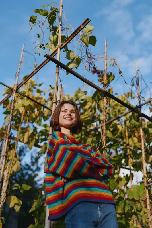 Woman smiling in a colorful striped sweater and casual outfit, young adult leaning against a wooden trellis in a lush garden outdoors, blue sky and joyful lifestyle portrait