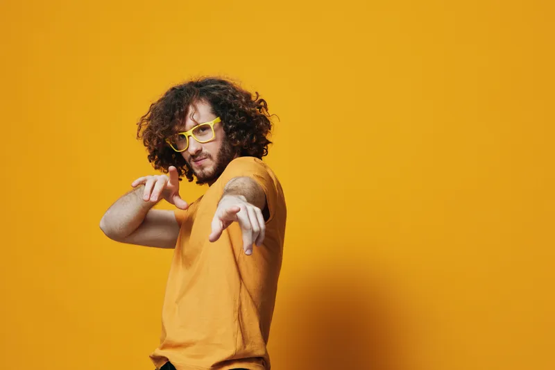 Young man with curly hair in a yellow shirt and glasses striking a playful pose against a vibrant yellow background, conveying confidence and creativity