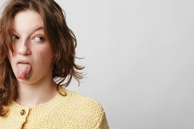 Young woman with brown hair wearing yellow sweater sticks out tongue and looks sideways on grey background. Concept of playful expression, fun, and casual attitude in portrait photography.